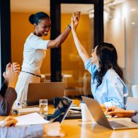 A group of coworkers gathers at a desk to celebrate a workplace achievement, as two women exchange a high-five surrounded by laptops and notes, fostering teamwork and positive engagement.